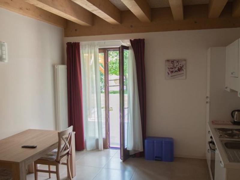 Soggiorno luminoso con cucina, tavolo da pranzo e porta finestra aperta verso l'esterno.