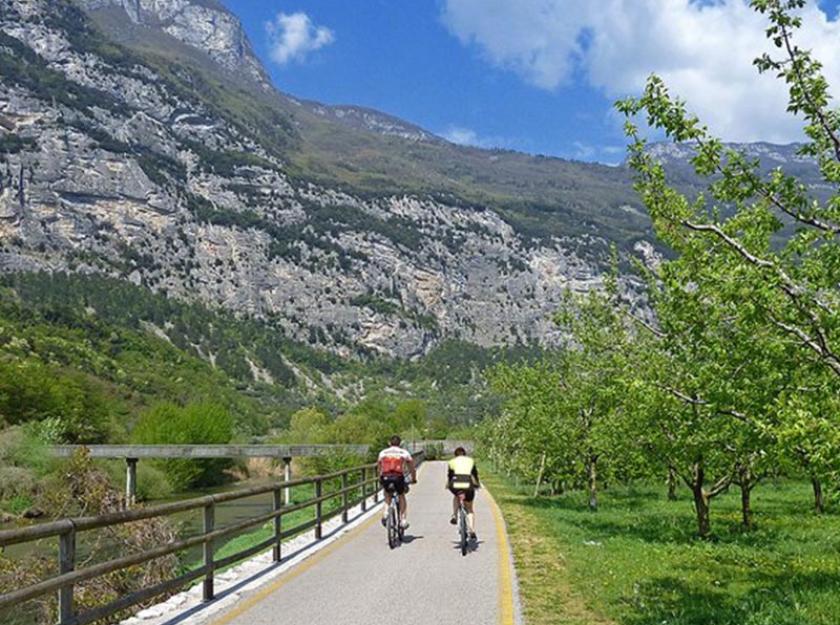 Ciclisti su pista ciclabile tra montagne e alberi in una giornata soleggiata.