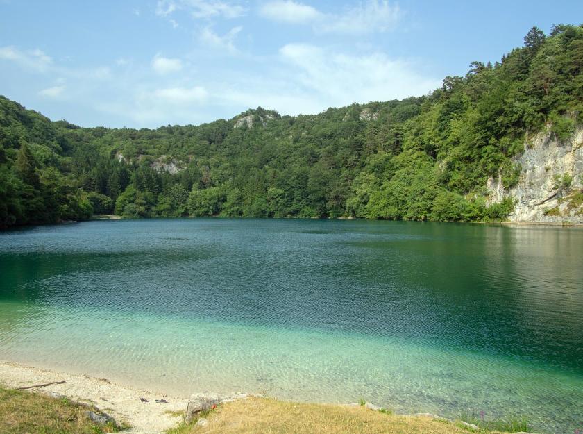 Lago circondato da foreste e colline, con acqua cristallina e cielo sereno.